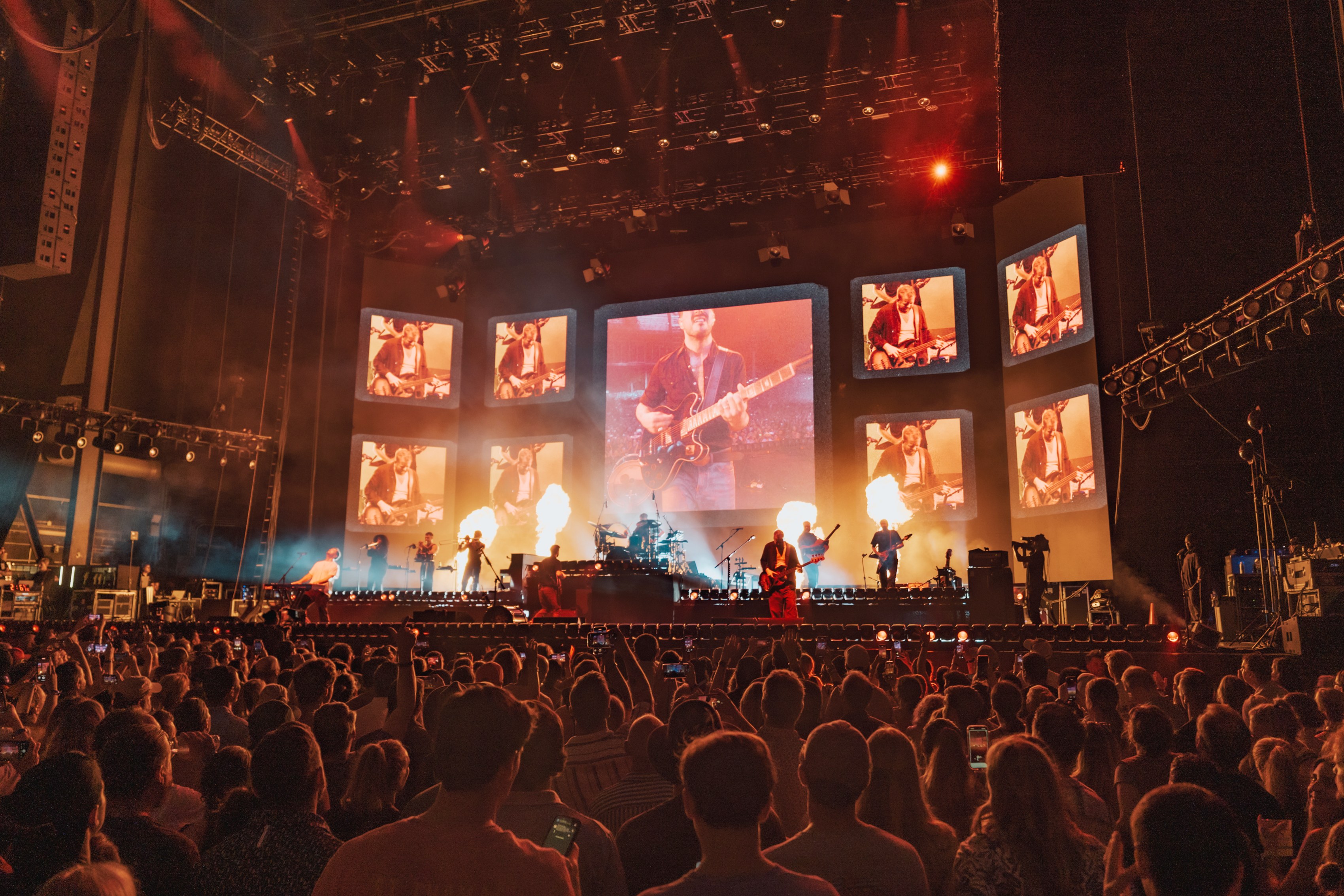A stage under a grid of screens shaped like old television sets, each carrying a close shot of the band, flame jets either side and the crowd lit orange in front